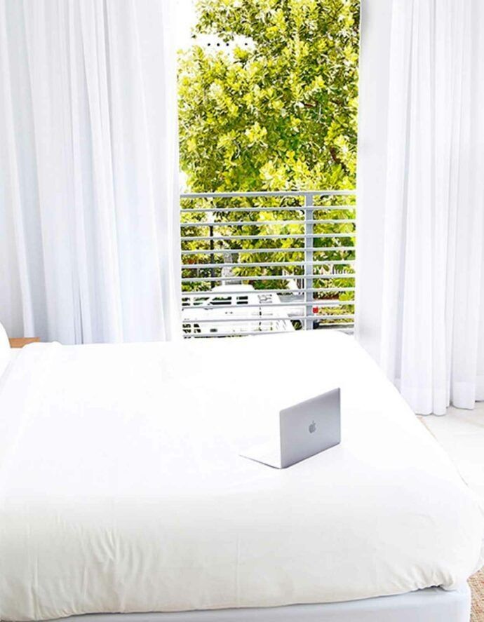 Laptop on a white bed, open in a bright room with window and curtains; view of a tree outside.