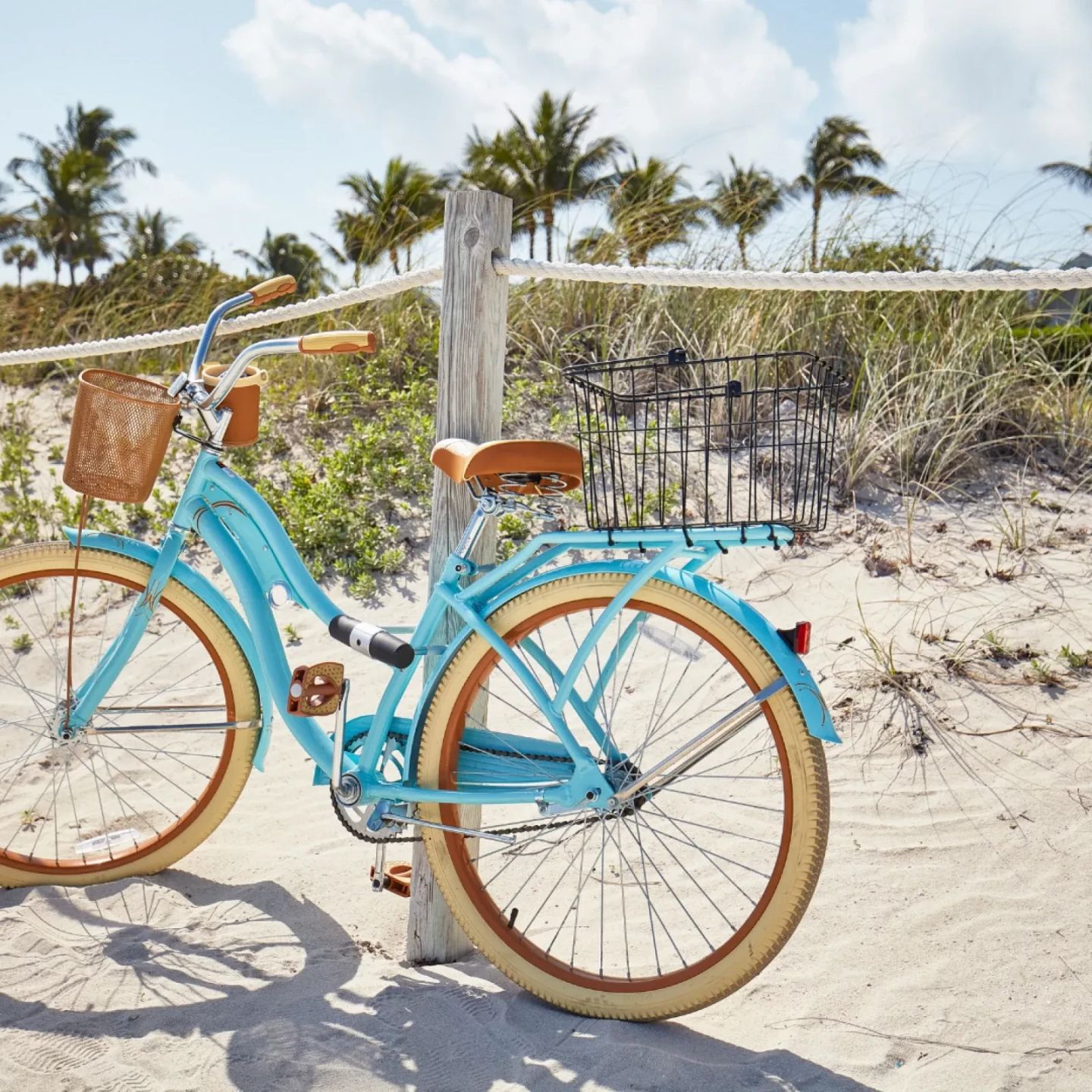 Light blue bicycle parked on a sandy beach, with a wooden fence and palm trees in the background.